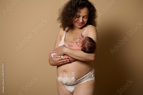 Authentic mom in underwear, smiles looking at her newborn baby she's holding, shows her body with postpartum scars flaws photo