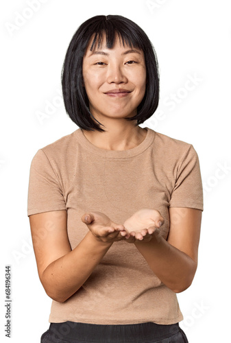 Young Chinese woman posing on studio background holding something with palms, offering to camera. © Asier