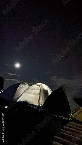 Camping under the moonlight, with a tent pitched and a campfire glowing in the middle of the night. photo