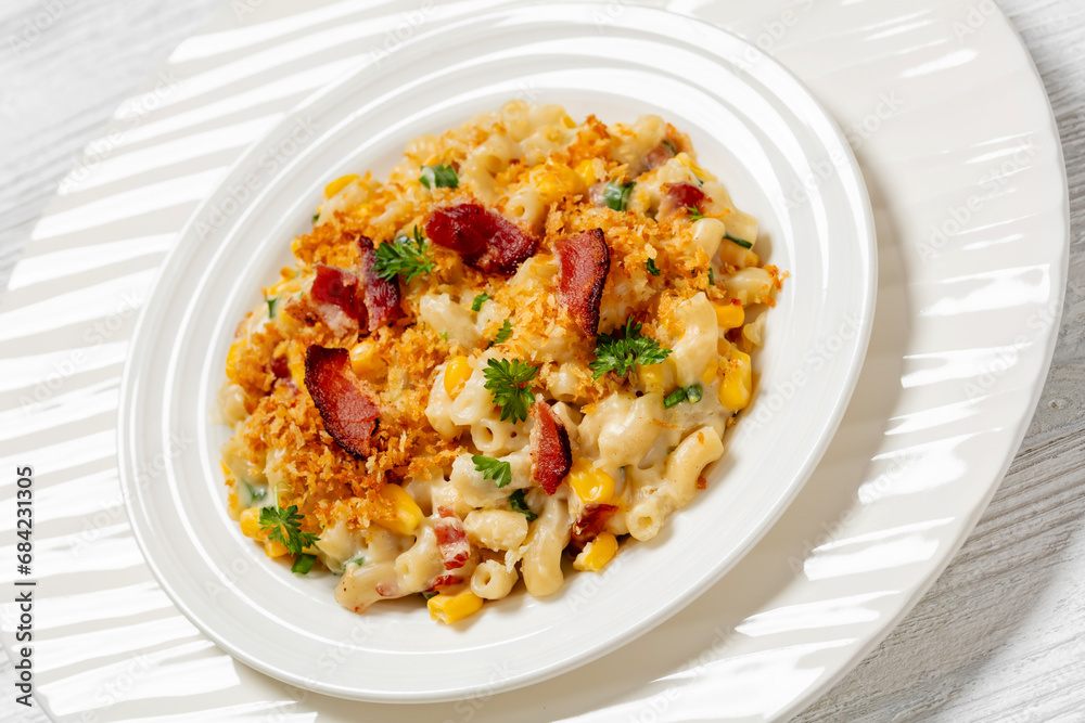 portion of mac and cheese with corn, bacon topped with panko breadcrumbs on white plate on white wooden table, dutch angle view, close-up portion of mac and cheese with corn, bacon topped with panko breadcrumbs on white plate on white wooden table, dutch angle view, close-up
