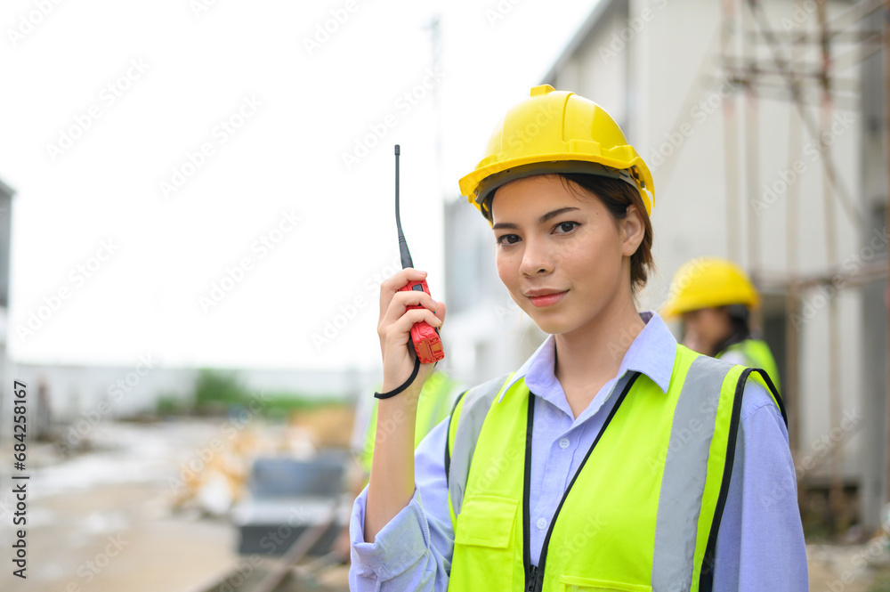 Young attractive construction woman in safety vest with yellow helmet working with radio, standing on building construction site. Home building project. Young attractive construction woman in safety vest with yellow helmet working with radio, standing on building construction site. Home building project.