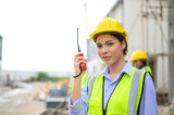 Young attractive construction woman in safety vest with yellow helmet working with radio, standing on building construction site. Home building project.