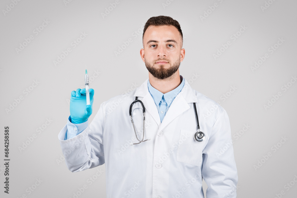 Serious young male doctor with syringe, white coat, gloves, grey background Serious young male doctor with syringe, white coat, gloves, grey background