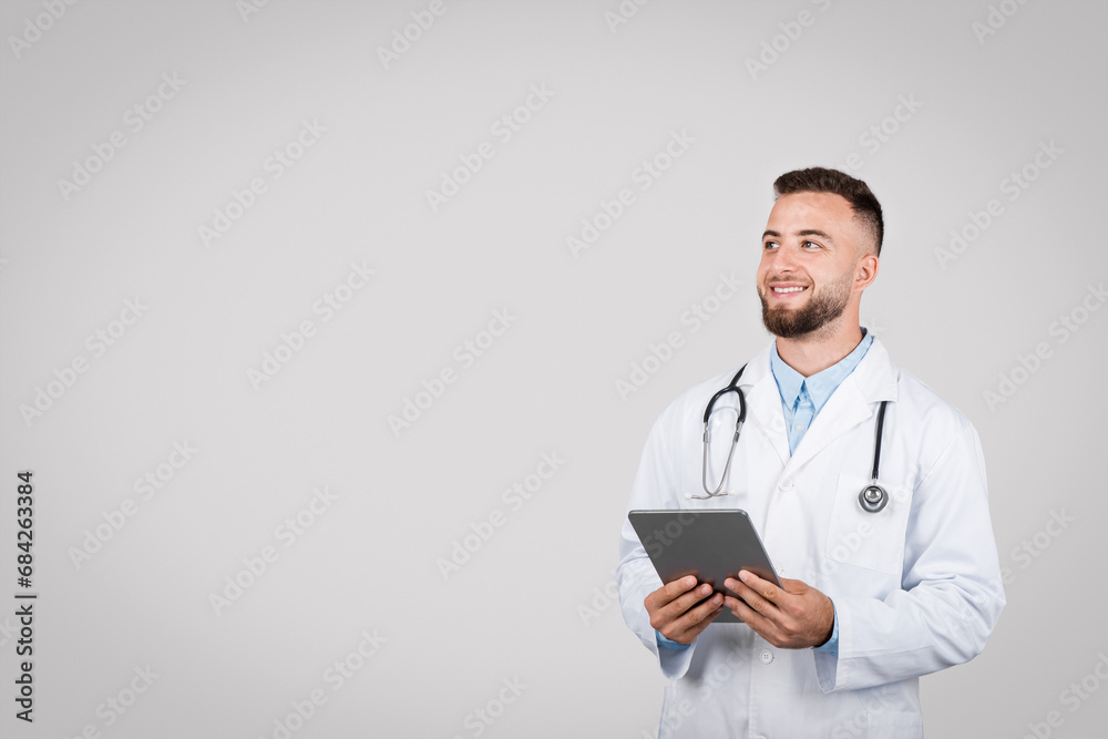 Young male doctor with tablet looking away on grey background Young male doctor with tablet looking away on grey background
