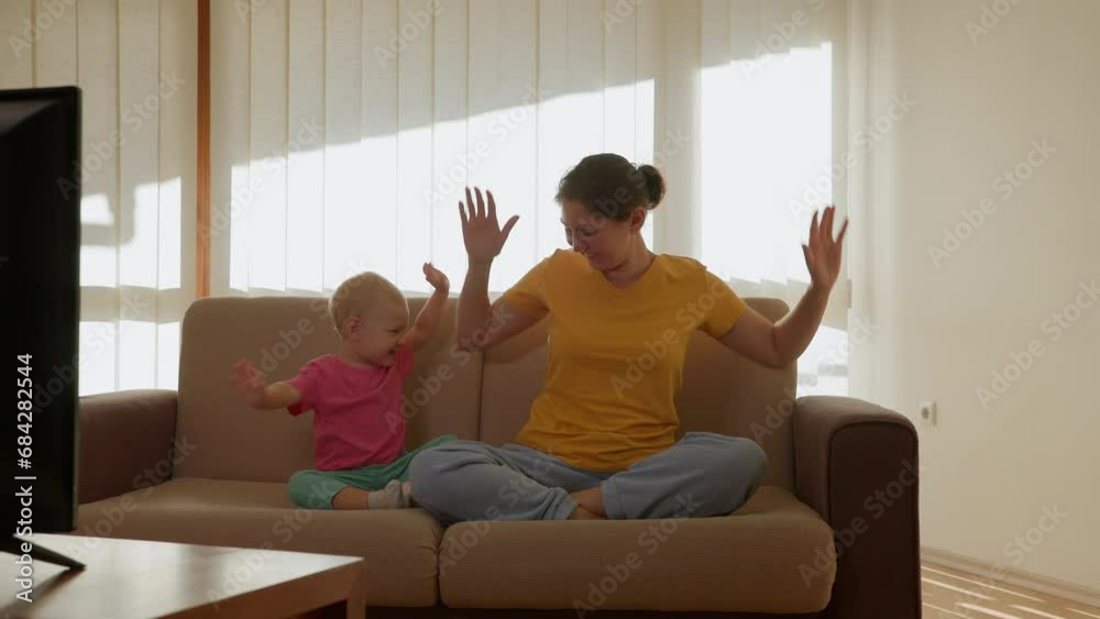 Mom and daughter are sitting on the sofa in front of the TV and doing exercises and having fun. High quality 4k footage Mom and daughter are sitting on the sofa in front of the TV and doing exercises and having fun. High quality 4k footage