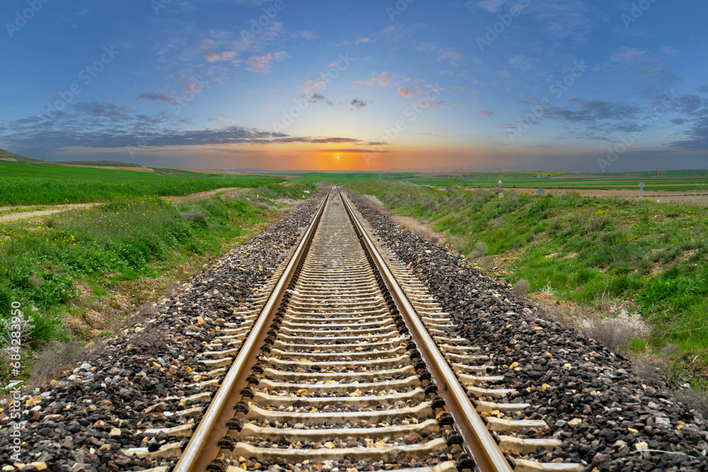 Railway stretching through greenery. Setting sun, sunset with blue cloudy sky landscape in the background Railway stretching through greenery. Setting sun, sunset with blue cloudy sky landscape in the background