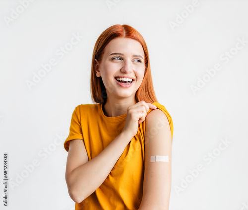 Portrait of a red-haired smiling young woman after getting a vaccine standing on a white background, showing her arm with bandage after receiving vaccination