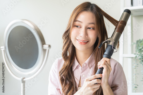 Beautiful curly brunette, attractive asian young woman, girl smiling using curler, curling iron long hair with ceramic curler straightener at home. Hairstyle and Hairdressing. Beauty and hair care.