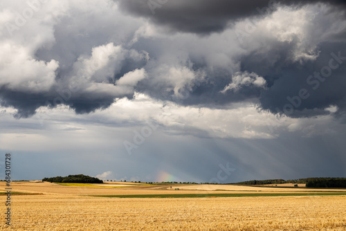 Heavy stormy clouds on sky with rainbow on background and harvester on field