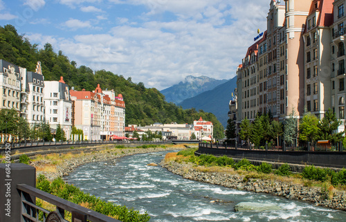 Obraz na płótnie Krasnaya polyana, Krasnodar Krai, Russia - July, 29, 2023: the embankment of the Mzymta River and modern architecture with luxury apartments among green hills on Krasnaya Polyana on a sunny summer day w salonie