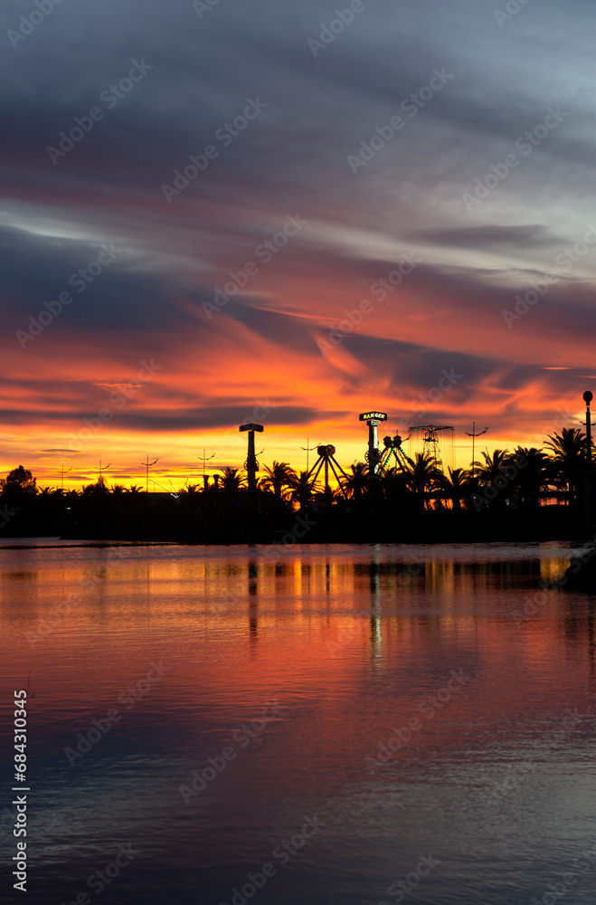 Dramatic sunset. Yellow, orange and red evening sky. Silhouettes of palm trees and park amusements