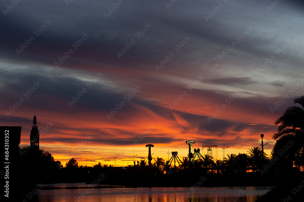 Dramatic sunset. Yellow, orange and red evening sky. Silhouettes of palm trees and park amusements