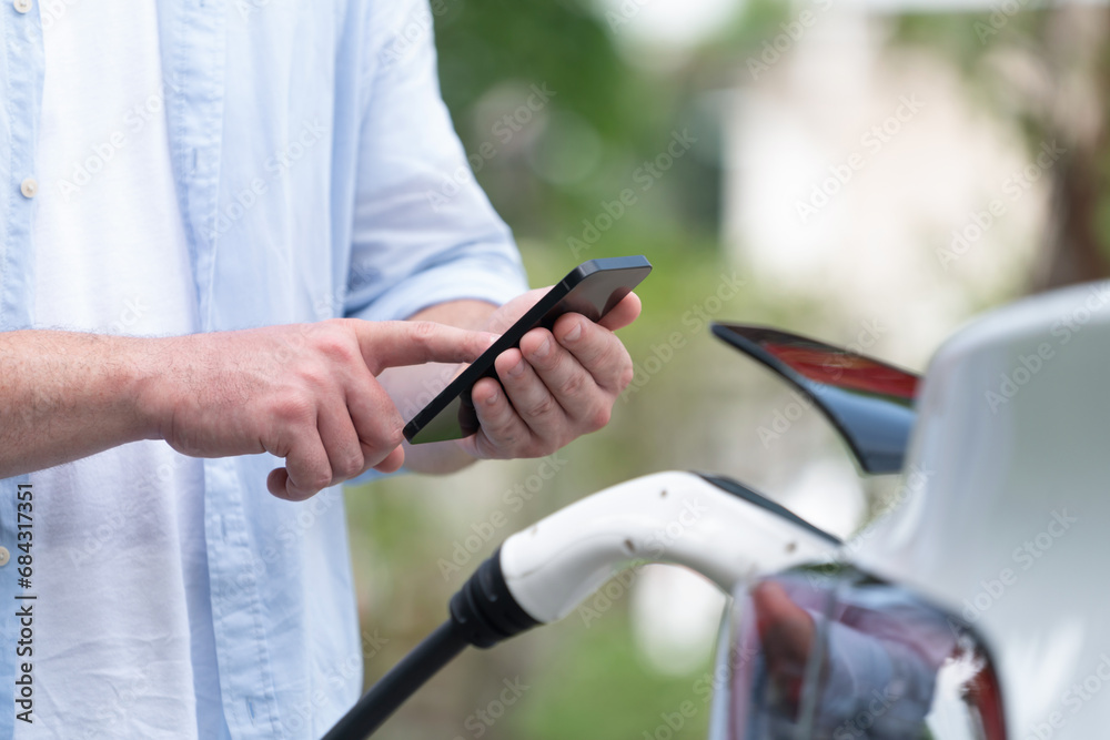 Modern eco-friendly man recharging electric vehicle from EV charging station, using Innovative EV technology utilization for tracking energy usage to optimize battery charging on smartphone.Synchronos