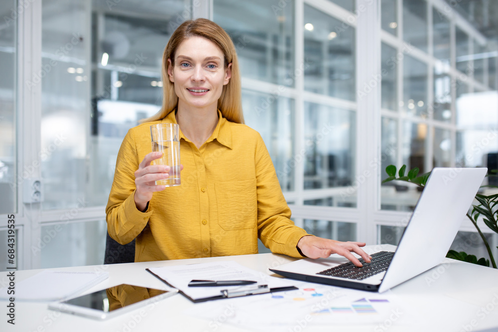 Portrait of a young woman in a yellow shirt working in the office at a laptop and drinking water from a glass, smiling and looking at the camera. Portrait of a young woman in a yellow shirt working in the office at a laptop and drinking water from a glass, smiling and looking at the camera.