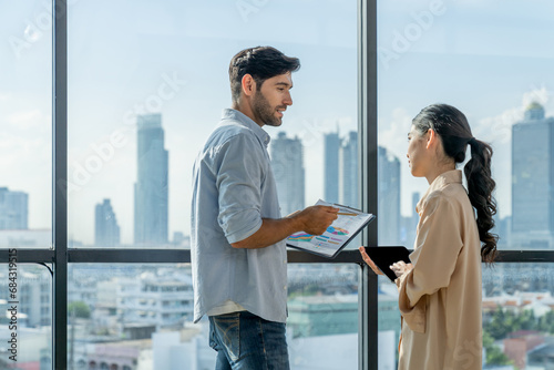 Professional businessman present financial statistic to investor while standing near panoramic window shows skyscraper or city view. Diverse businesspeople discuss about marketing plan. Tracery.