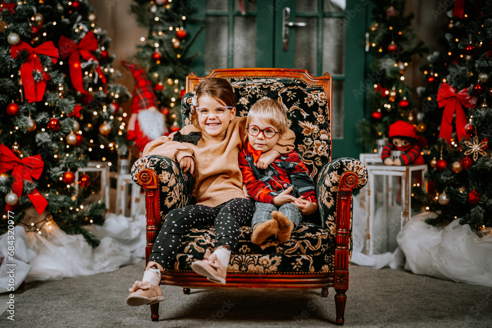 A child girl hugs her younger brother while sitting on a chair against the backdrop of a New Year tree and Christmas gifts. Little children brother and sister having fun during the Christmas holidays