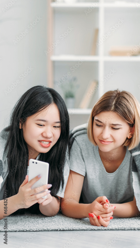 Device leisure. Technology youth. Two friends girls enjoying smartphone gadget internet social media content fun resting in light interior on floor at home. Device leisure. Technology youth. Two friends girls enjoying smartphone gadget internet social media content fun resting in light interior on floor at home.