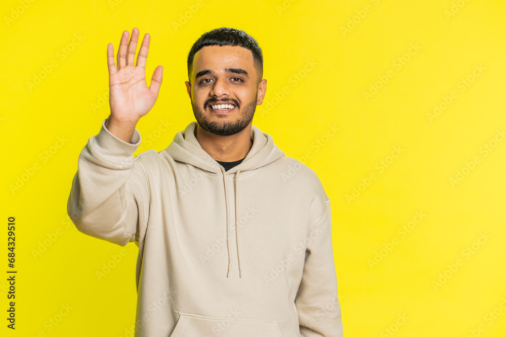 Lovely Indian man smiling friendly at camera, waving hands gesturing hello greeting or goodbye welcoming with invitation hospitable expression. Arabian Hindu guy isolated on yellow studio background Lovely Indian man smiling friendly at camera, waving hands gesturing hello greeting or goodbye welcoming with invitation hospitable expression. Arabian Hindu guy isolated on yellow studio background