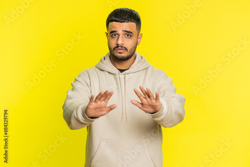 Hey you, be careful. Strict Indian man warning with admonishing hands gesture, saying no, be careful, scolding and giving advice to avoid danger, disapproval sign. Guy on yellow studio background photo