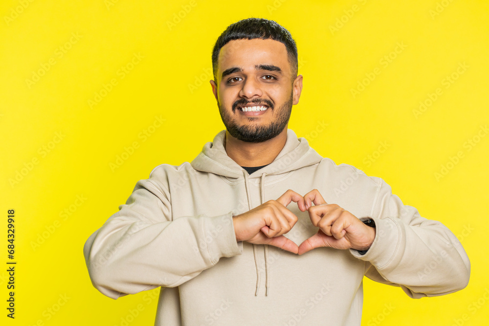 Man in love. Smiling attractive Indian man makes heart gesture demonstrates love sign expresses good positive feelings and sympathy. Handsome Arabian Hindu young guy isolated on yellow wall background Man in love. Smiling attractive Indian man makes heart gesture demonstrates love sign expresses good positive feelings and sympathy. Handsome Arabian Hindu young guy isolated on yellow wall background