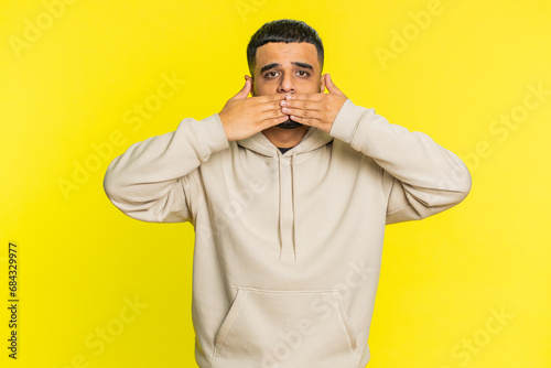 I will not say anything. Frightened Indian man closing mouth with hands, looking intimidated scared at camera gestures no, refusing to tell terrible secret unbelievable truth. Guy on yellow background