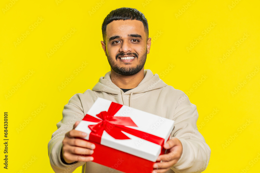 Happy smiling Indian man presenting birthday gift box stretches out hands, offer wrapped present career bonus, celebrating party, promotion discount sale. Arabian guy isolated on yellow background Happy smiling Indian man presenting birthday gift box stretches out hands, offer wrapped present career bonus, celebrating party, promotion discount sale. Arabian guy isolated on yellow background