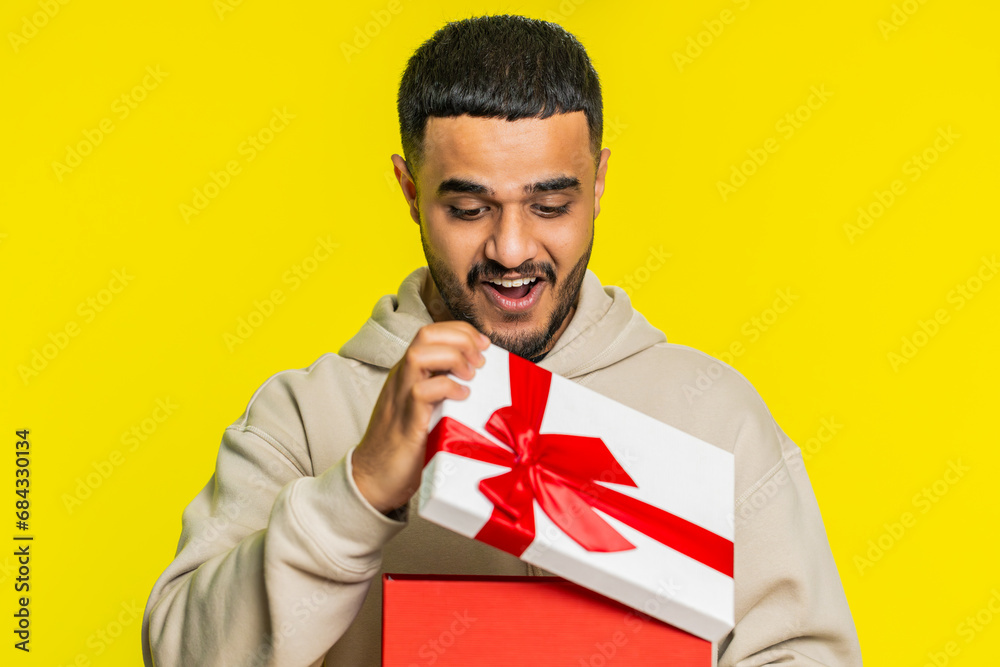 Happy Indian young man opening birthday gift box with red ribbon. Holidays surprise concept. Smiling Arabian Hindu guy receive great wrapped present celebrating isolated on yellow studio background