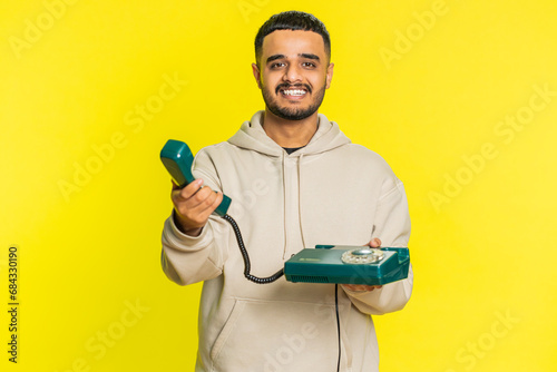 Hey you, call me back. Indian young man talking on wired landline vintage telephone of 80s, advertising proposition of conversation, online shopping, hotline. Arabian guy isolated on yellow background