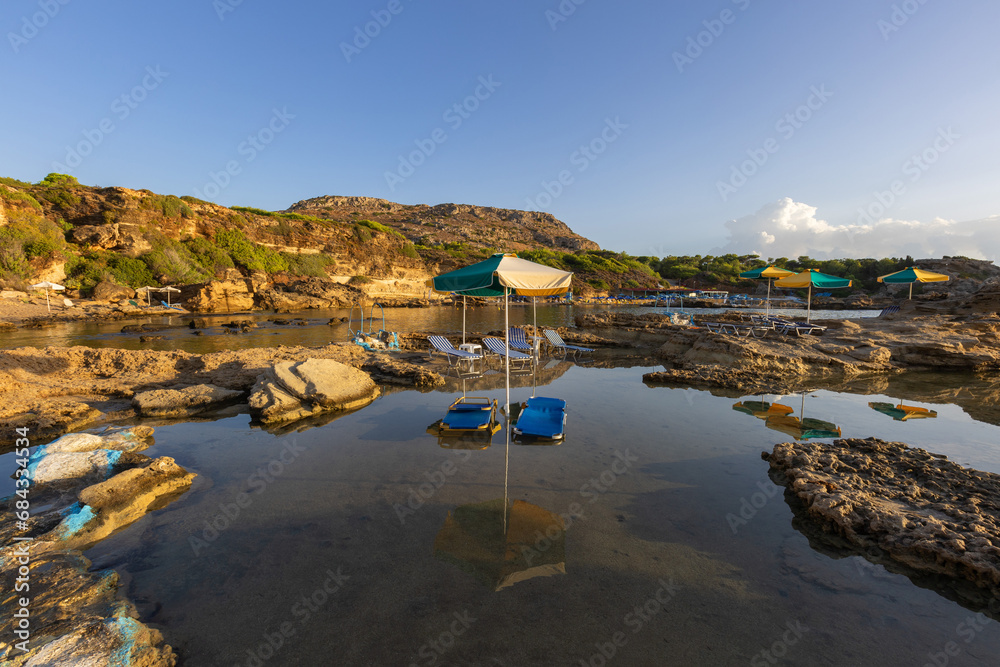 A picturesque bay on the island of Rhodes with shallow water that forms small lagoons between the rocks. There are sunbeds and umbrellas in the water.