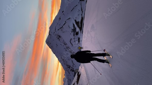Two skiers smoothly accelerate along a perfectly prepared track at dawn. There is no one at the ski resort yet. Sun has not yet come out from behind the mountain. Golden light reflects off the clouds