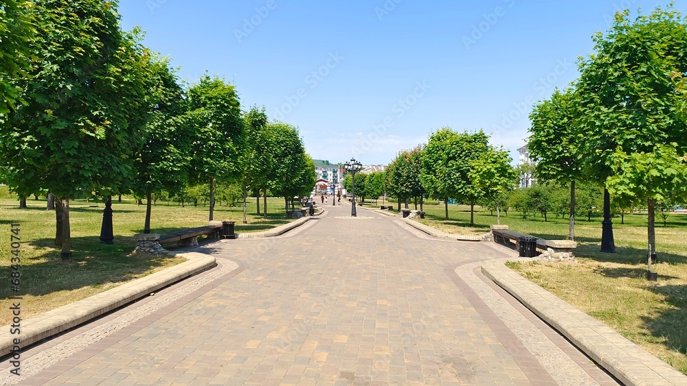 In the city park, paving stone walkways with curbs are laid among mowed lawns and various trees. There are metal poles with lanterns on them. Behind the park are buildings. Sunny summer weather