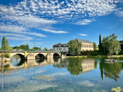 view of a stone bridge in a small French town over a river on a summer day with some white clouds
