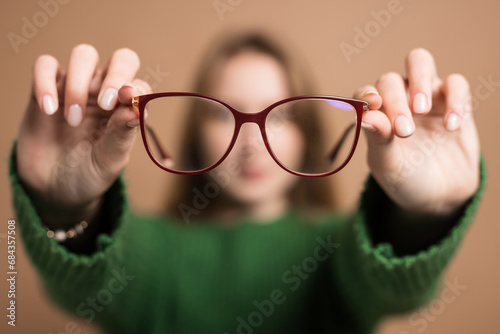 Poor vision concept, Young girl with poor vision holds glasses with hands forward on beige background photo