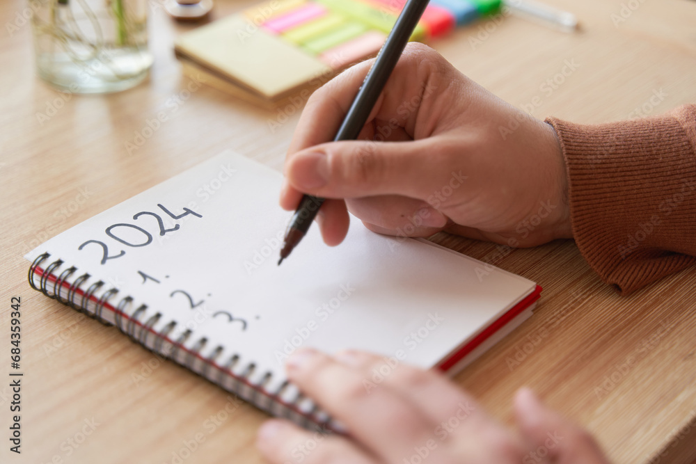 Young hands writing in notebook 2024 new year's resolutions index list over wooden table Young hands writing in notebook 2024 new year's resolutions index list over wooden table