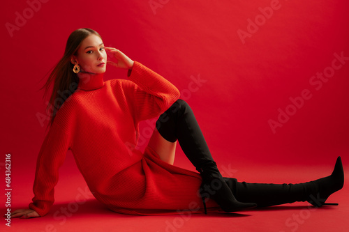 Fashionable confident woman wearing trendy red long knitted turtleneck dress, over the knee high heel boots, posing on red background. Full-length studio fashion portrait. Copy, empty space for text photo