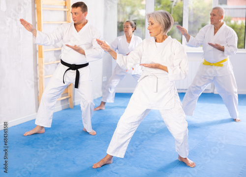 Diligent old man attendee of karate classes practicing kata standing in row with others in sports hall