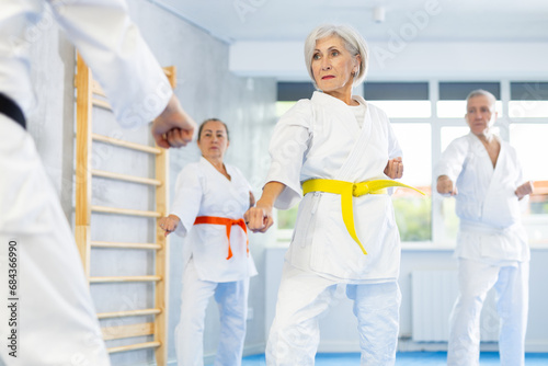 Diligent old man attendee of karate classes practicing kata standing in row with others in sports hall