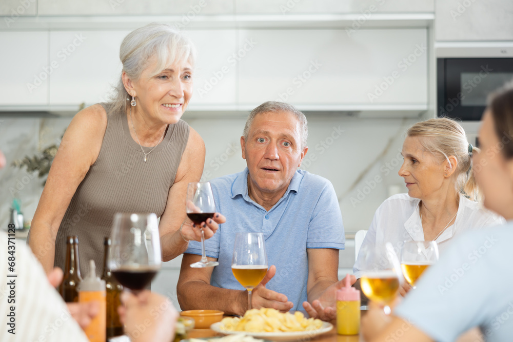 Happy carefree older man and women gathering around kitchen table, sipping alcoholic drinks with snacks, cheerfully chatting, laughing and sharing stories. Seniors communication concept