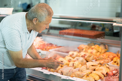Adult man buyer in casual clothes chooses raw chicken meat in butcher shop