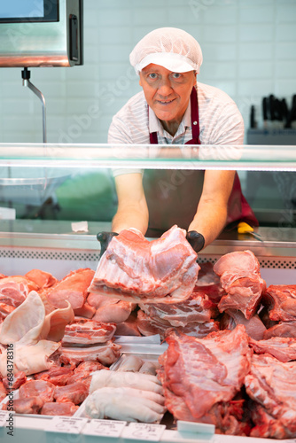 In local butcher shop senior male seller in apron and gloves takes out large raw pork boneless meat from glass showcase of refrigerator. Sales assistant shows ribbon damp gammon of bacon in meat store
