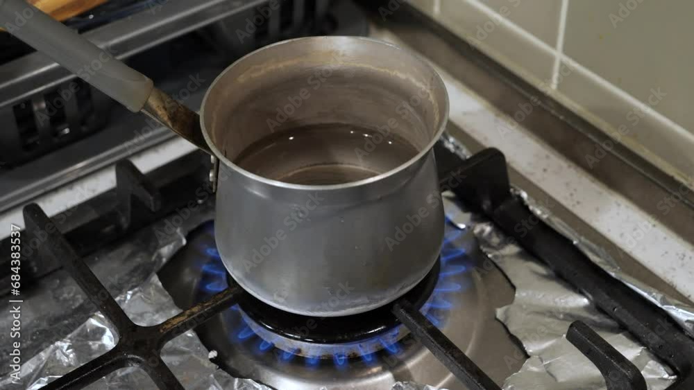 Boiling water with the Turkish coffee warmer on the gas stove. Closeup orbit shot of the metal pot utensil with handle, half-filled with water, standing on the stovetop covered by silver foil paper. Boiling water with the Turkish coffee warmer on the gas stove. Closeup orbit shot of the metal pot utensil with handle, half-filled with water, standing on the stovetop covered by silver foil paper.