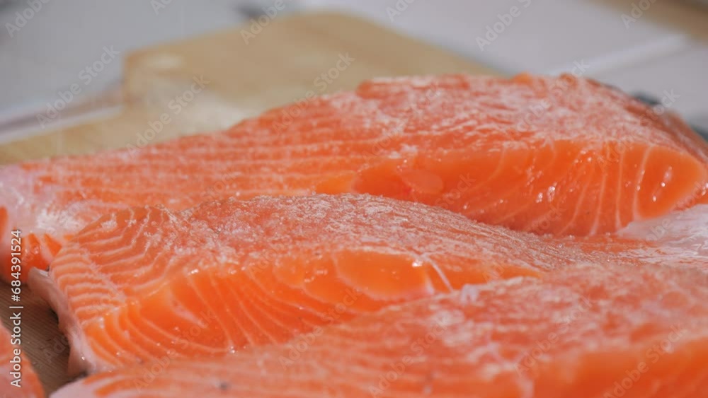 A piece of fresh red fish is sprinkled with salt in slow motion close up view. Seasoning salmon steak. Chef adding spices on raw steak before baking A piece of fresh red fish is sprinkled with salt in slow motion close up view. Seasoning salmon steak. Chef adding spices on raw steak before baking
