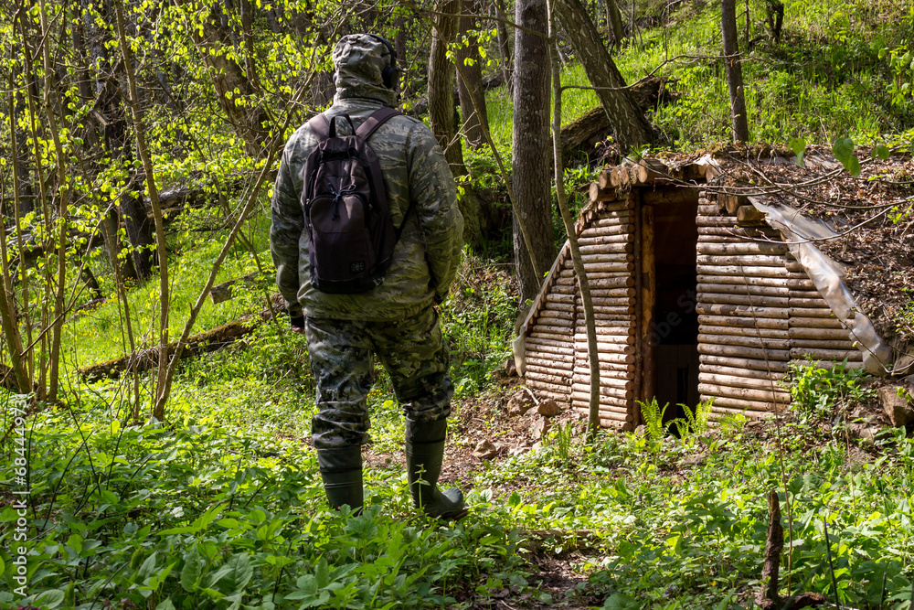 A man in a camouflage suit with a backpack from his back against the background of a forest dugout built in a wild forest