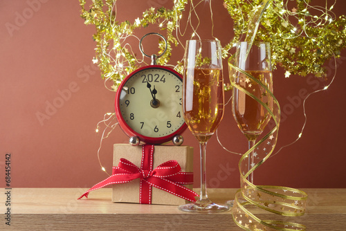 New Year 2024 celebration festive background with champagne glasses, gift box and clock on wooden table over golden decorations lights
