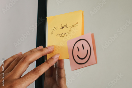 Woman putting a sticky note with phrase You Look Great Today! on the mirror.