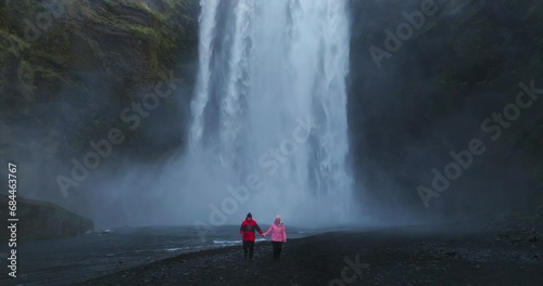 Tourist Couple With Skogafoss Waterfall In The Background In Iceland. Zoom Out