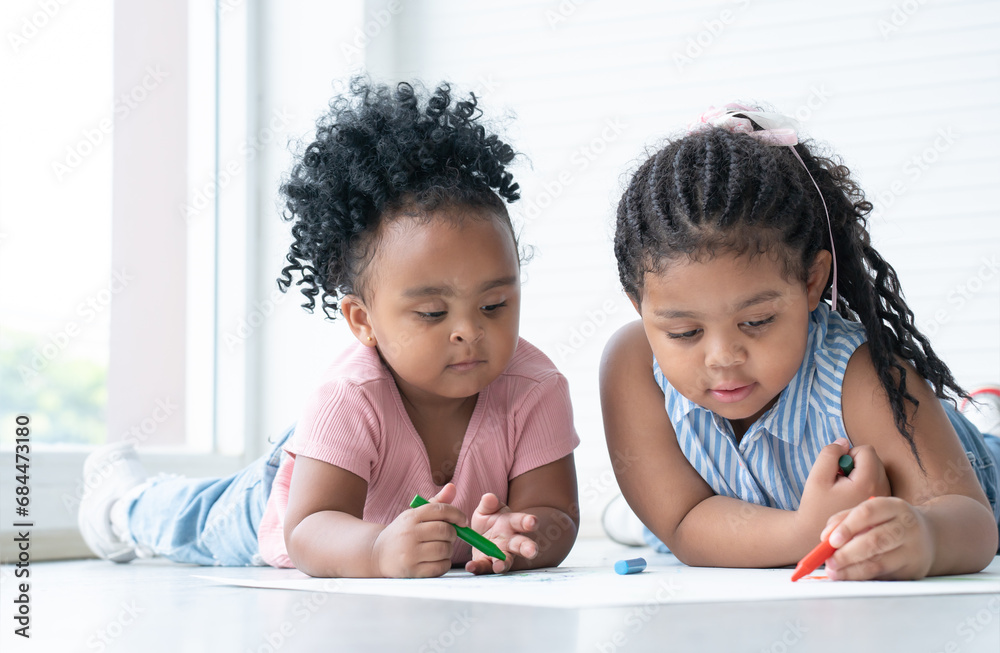 Fototapeta premium African cute little kid girl teaching her sister drawing on paper while lying on the floor. Adorable children have fun drawing with colorful crayons at home. Education, family lifestyles, creativity
