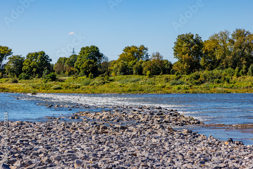 Abundant gravel and stones in Maas river, flowing water, alluvial terrain, Belgian countryside with trees in background, sunny day, blue sky in Maasvallei nature reserve in Meers, Elsloo, Netherlands