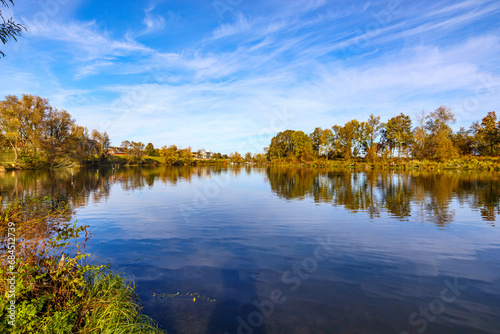Landscape of a river surrounded by autumn trees against blue sky in background, mirror reflection on water surface, Belgian nature reserve De Wissen Maasvallei, sunny day in Dilsen-Stokkem, Belgium