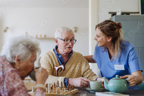 Happy healthcare worker serving tea to senior couple at table photo